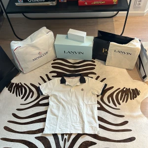 Vit Moncler piké med marinblå krage - Vit pikétröja från Moncler med klassisk marinblå krage och matchande detaljer på ärmslut. Tröjan har korta ärmar, broderad Moncler-logga på bröstet och snygga röda ränder på insidan av kragen. Materialet är mjuk bomull som känns skönt mot huden. Helt ÄKTA köpt på Qatar flygplats.