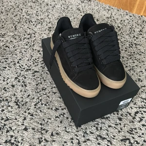 Svarta  Numeris med beige sula - Stilrena svarta sneakers från Numeris med breda svarta snören och kontrasterande beige sula. Skorna har en rund tå och är tillverkade i mocka för en lyxig känsla. Perfekta för dig som gillar minimalistisk och modern streetstyle.