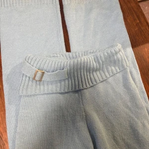 Ljusblå stickade mjukisbyxor - Säljer ett par ljusblå stickade mjukisbyxor med ribbad midja och dekorativt bälte med metallspänne. Byxorna har en avslappnad passform och är perfekta för chill dagar hemma. Materialet är mjukt och skönt mot huden.