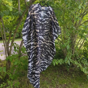 Zebramönstrad långklänning i polyester - Flowy långklänning med zebramönster i svart, vit och beige. Klänningen har v-ringad hals med snörning och är tillverkad i ett lätt och svalt polyestertyg. Perfekt för dig som gillar statement-plagg med loose passform och coola mönster.  Köpte den här på vinted men den har bara blivit hängandes då den är alldeles för lång för mig som är 167cm lång. Defekt finns då den är sydd inuti, så kolla bilderna innan köp, jag tar ej emot returer.  Katter finns i hemmet.