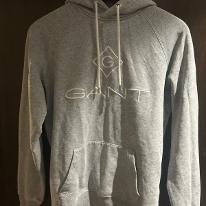 Grå hoodie från GANT med broderad logga - Snygg grå hoodie från GANT med broderad logga framtill. Klassisk modell med huva, snörning och känguruficka. Mjuk och skön i bomullsmix, perfekt för chill dagar. Långärmad och med ribbade muddar för extra komfort.