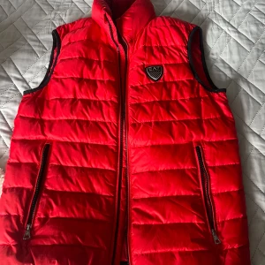 Röd dunväst från EA7 Emporio Armani - Snygg röd dunväst från EA7 Emporio Armani med quiltad design och svart kant vid ärmhålen. Västen har två fickor med dragkedja och ett EA7-märke på bröstet. Perfekt att slänga över en hoodie för en sportig look.