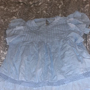 Ljusblå blus med volangärmar - Supersöt ljusblå blus med volangärmar och diskret prickigt mönster. Blusen har en rund halsringning, smockad överdel och spetsdetalj längst ner. Perfekt för dig som gillar romantisk och feminin stil.