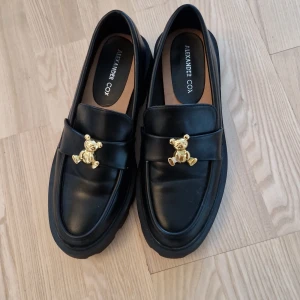 Svarta loafers Alexander Cox med björn - Oanvända snygga svarta loafers från Alexander Cox i skinn med chunky platt sula och rund tå. På ovansidan sitter en cool guldfärgad björn-detalj som ger skorna en unik vibe. Perfekta för dig som gillar att sticka ut med din stil.