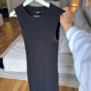 Svart långärmlös klänning från Bik Bok - Säljer en svart, ribbad långklänning med slits från Bik Bok i storlek XS. Klänningen är ärmlös med rund halsringning och har en slim passform som sitter snyggt mot kroppen. Perfekt för dig som gillar minimalistisk stil och vill ha en enkel men stilren look. 