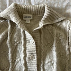 Beige stickad kofta från Monki - Supermysig beige stickad kofta från Monki med bred krage och stora vita knappar framtill. Koftan har ett unikt upphöjt blommönster och ribbade detaljer vid krage, knappslå och nederkant. Perfekt för lager-på-lager och chill dagar.