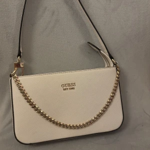 Vit axelväska från Guess med kedja - Snygg vit axelväska från Guess med guldfärgad kedjedetalj och logga framtill. Väskan har struktur i materialet, dragkedja på baksidan och justerbart axelband. Perfekt storlek för det viktigaste och ger en clean look till outfiten.