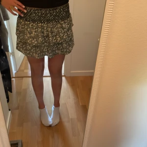 Blommig kortkjol från Zara - Supersöt kortkjol från Zara med svart och vit blommigt mönster. Kjolen har smockad midja och volangkant som ger en flowy och chill vibe. Perfekt för dig som gillar att sticka ut med detaljer och mönster. Passar dig som vill ha en bekväm och snygg kjol till sommaren. Med inbyggda shorts 