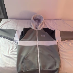 Grå och vit hoodiejacka från McKenzie - Snygg hoodiejacka från McKenzie i grått, vitt och svart. Jackan har dragkedja framtill, huva och två fickor. Perfekt för dig som gillar en sportig och clean stil. Materialet känns mjukt och bekvämt, och färgblocken ger en modern vibe.