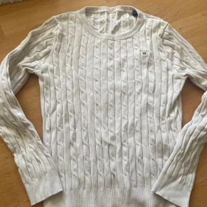 Snygg tröja från GANT - Snygg vit kabelstickad tröja från GANT med rund hals och broderad logga på bröstet. Tröjan är i storlek 158/164. Fint skick och knappt använd, inga fläckar eller missfärgningar.