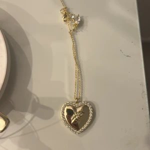 Guldigt halsband med hjärthänge från ioku - Snyggt halsband i guld med ett stort hjärthänge som är dekorerat med små pärlor runt kanten. Kedjan är tunn och elegant, perfekt för att ge din outfit lite extra bling. Kommer i originalask från Looki.