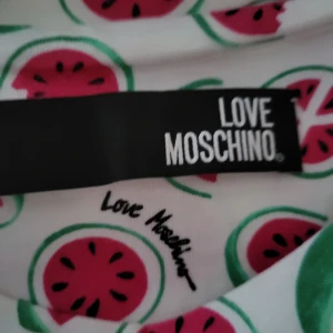 Love Moschino klänning med vattenmelon - Säljer en vit klänning från Love Moschino med färgglatt vattenmelonmönster över hela plagget. Klänningen har rund hals och korta ärmar, och är gjord i mjuk bomull. Perfekt för dig som gillar lekfulla prints och vill sticka ut med en unik design.