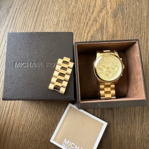 Guldig Michael Kors klocka i stål - Snygg guldig klocka från Michael Kors med länkarmband i rostfritt stål. Något använd, därav vissa små skavanker (se sista bilden). Fungerar som den ska. Originalförpackning och extra länkar medföljer.