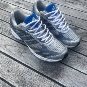  Adidas sneakers med blå detaljer - Snygga Adidas sneakers i silver och blått med klassiska tre ränder på sidan. Ovandelen är i ventilerande mesh och syntet, med vita snören och vit sula. Perfekta för dig som gillar sportig stil och vill sticka ut med retrovibbar.
