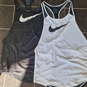 Nike Dri-Fit linnen svart & vitt - Två sportiga linnen från Nike Dri-Fit, ett svart med vitt Swoosh-tryck och ett vitt med svart Swoosh. Båda har racerback och är tillverkade i ett lätt, svalt material som passar perfekt för träning. Snygg och enkel design med hög halsringning.