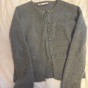 Grå stickad kofta från Zara - Mysig grå stickad kofta från Zara med rund hals och pärlknappar framtill. Koftan har långa ärmar och en enkel, klassisk design som är lätt att matcha med olika outfits. Perfekt för kyliga dagar och passar dig som gillar stilrena plagg.