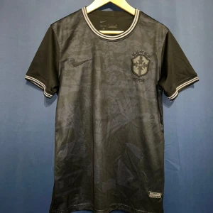 Svart Brasil Nike fotbollströja - Snygg svart Brasil fotbollströja från Nike med diskret mönster och broderat CBF-emblem på bröstet. Tröjan har rund hals, korta ärmar och vita ränder på muddar och krage. Materialet är lätt och andas, perfekt för match eller träning.