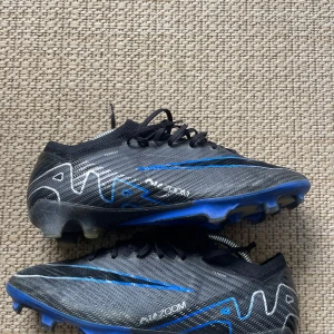 Nike Mercurial Air Zoom fotbollsskor - Snygga Nike Mercurial Air Zoom fotbollsskor i svart och blått med silverdetaljer. Skorna har snörning, låg profil och mönstrad ovandel i syntetmaterial. Sulan är blå med fasta dobbar för gräsplan. Perfekta för dig som vill ha snabbhet och kontroll på planen.