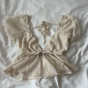 Beige omlottopp med knyt från SHEIN - Supersöt beige omlottopp från SHEIN med korta puffärmar och knyt både i ryggen och framtill. Tunt, lätt skimrande tyg med struktur och v-ringning. Perfekt för dig som gillar romantisk och feminin stil.