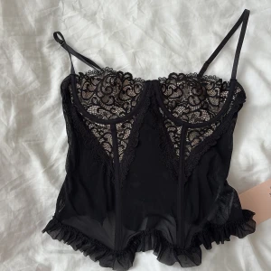 Svart spetskorsett med volang - Supersnygg svart korsett med tunna axelband, spetsdetaljer över bysten och volangkant nedtill. Toppen har meshmaterial och dragkedja i ryggen för enkel på- och avtagning. Perfekt för dig som vill ha en edgy och feminin look.