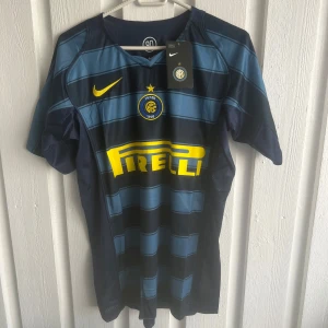 Inter Adriano 10 Nike fotbollströja - Snygg Inter fotbollströja från Nike med Adriano 10 på ryggen. Tröjan är blå med mörkare blå ränder och gula detaljer, inklusive Pirelli-loggan och klubbmärket. Kortärmad modell i lätt och ventilerande material, perfekt för match eller träning.