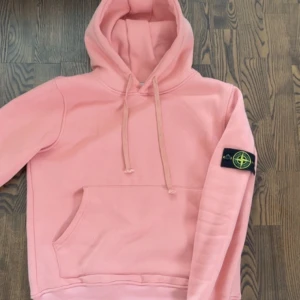 Rosa hoodie från Stone Island - Säljer en rosa hoodie från Stone Island med klassisk patch på ärmen. Tröjan har huva med snörning och en stor magficka framtill. Mjuk bomullskvalitet och relaxed fit gör den perfekt för chill dagar. Ikonisk streetwear-vibe och snygg färg som sticker ut.