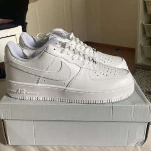 Nike Air Force 1 helvita sneakers - Klassiska Nike Air Force 1 i helvitt skinn med perforerad tå, vit sula och snörning. Snygg och tidlös modell med låg profil och ikonisk Swoosh-logga på sidorna. Perfekta för dig som vill ha en clean och fräsch look.