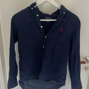 Mörkblå linneskjorta Polo Ralph Lauren - Hej! Jag säljer min begagnade mörkblå skjorta från Polo Ralph Lauren! Storleken är i barn(m10-12) men är stor i storleken och passar nån runt 160cm eller mindre. Jag är runt163,164 och den sitter ganska kort och är för liten för mig och därför säljer jag den. Skjortan köptes begagnat på Plick och inte används jättemycket sen jag fick pga för liten men den är i bra skick! Köptes för ungefär 500kr på Plick men nypris är runt 1000kr och mitt pris är 449 skriv om ni har några frågor!