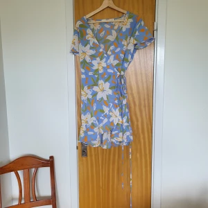 Blommig omlottklänning i blått - Superfin kort omlottklänning i blått med stora vita blommor med gröna och orange detaljer. Klänningen har v-ringning, korta ärmar och knyt i midjan. Perfekt för sommaren. Står storlek L på etiketten men passar storlek 36-38 och känns inte för stor. 