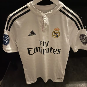 Real Madrid Ronaldo 7 matchtröja Adidas - Säljer en ikonisk vit Real Madrid fotbollströja från Adidas med svarta detaljer och tryck. Tröjan har korta ärmar, klubbmärke på bröstet, Champions League-märke på ärmen och Ronaldo 7-tryck på ryggen. Tillverkad i lätt och ventilerande climalite-material.