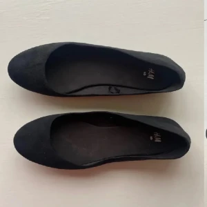 Svarta ballerinaskor från H&M - Klassiska svarta ballerinaskor från H&M i enkel och stilren design. Skorna har rund tå och platt sula, perfekta för en clean look. Tillverkade i ett mjukt syntetmaterial som känns bekvämt på foten.