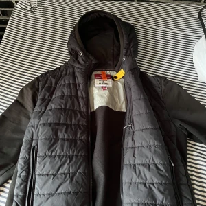 Svart cardigan från Parajumper - Snygg svart cardigan från Parajumpers med huva och coolt patch på ärmen. Jackan har dragkedja, quiltad framsida och detaljer i gult och orange. Materialmix av fleece och syntet ger både värme och stil. Perfekt för dig som vill hålla dig varm och snygg på sena sommarkvällar köpt på jhonells för 4000kr priset är ej hugget i sten. Storlek M men passar även S