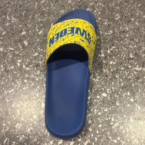 Blå och gula sandaler med SWEDEN-tryck - Snygga slides i blått med bred gul rem där det står SWEDEN i blått. Remmen har ett coolt stänkmönster och skon har en platt sula. Perfekta för sommaren eller stranden, och riktigt sköna att glida runt i.