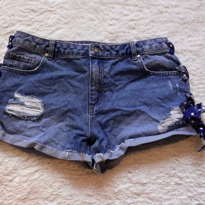 Blå jeansshorts med prickiga rosetter - Snygga blå jeansshorts med slitna detaljer och upprullad kant. Sidorna är dekorerade med marinblå band med vita prickar som är knutna till rosetter, vilket ger en unik och lekfull look. Perfekta för varma dagar när du vill sticka ut lite extra.