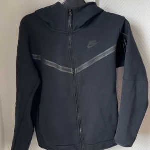 Svart Nike hoodiejacka med dragkedja - Svart hoodiejacka från Nike med hel dragkedja framtill och huva. Jackan har en diskret grå rand över bröstet och Nike-logga på vänster sida. Tillverkad i mjukt bomullsmaterial och har långa ärmar. Perfekt för en sportig och avslappnad stil.