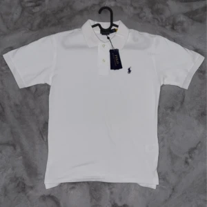 Vit pikétröja från Polo Ralph Lauren - Klassisk vit pikétröja från Polo Ralph Lauren med korta ärmar och broderad marinblå logga på bröstet. Tröjan har krage, knappar framtill och är tillverkad i mjuk bomull. Perfekt för en clean och stilren look.