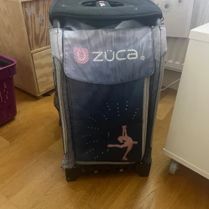 Züca resväska med konståkningsmotiv - Stor och rymlig resväska från Züca i grått och blått med rosa detaljer. Väskan har flera fack, robusta hjul som lyser när den rullar och dragkedjor. Framsidan pryds av ett konståkningsmotiv med glittrande stenar. Perfekt för att få med sig allt till träning eller tävling. Lagom med plats till allt man behöver för att träna.
