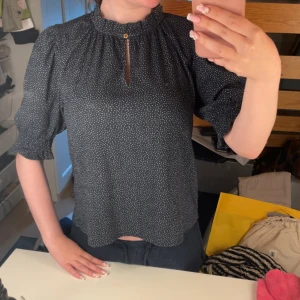 Svart prickig blus med volangkrage - Supersöt svart blus med små vita prickar, volangkrage och en liten knappdetalj vid halsen. Blusen har puffiga halvlånga ärmar med resår och en lätt avslappnad passform. Perfekt för dig som gillar en söt och trendig look.