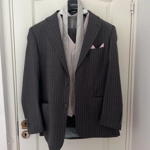 Randig svart kavaj från cavaliere ny pris 8500 - Snygg svart kavaj med tunna vita ränder från cavaliere . Klassisk passform med två knappar framtill och tre knappar vid ärmslut. Matchande väst i ljusrosa med diskret mönster och grå slips ingår. Perfekt för dig som vill sticka ut med stil.Med kostym byxor och slips 