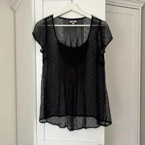 Svart transparent blus från Noa Noa - Supersöt svart transparent blus från Noa Noa i luftig modell. Blusen har korta ärmar, broderad detalj vid halsen och är dekorerad med tunna vertikala ränder och små prickar. Perfekt att styla lager på lager för en cool look.