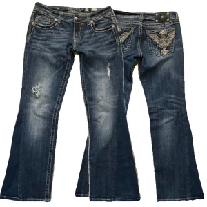 Blå bootcut Miss Me jeans med broderad detaljer - Snygga blå bootcut jeans från Miss Me med unika broderade detaljer på bakfickorna. Jeansen har en cool sliten look och är perfekta för en avslappnad stil. De har en lågmidjad passform och är dekorerade med knappar och dragkedja.