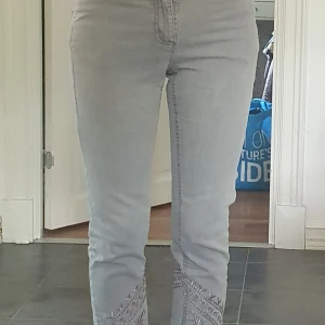 Grå jeans med broderi och dragkedja -  Ljusgrå jeans med broderade blommor och cirklar nertill på benen. Byxorna har dragkedja vid benslut och är i croppad modell. Ca 35 cm tvärs över vid midjan.