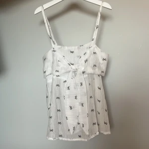 Vit linnetopp med knytning och svarta rosetter - Supersöt vit linnetopp med svarta rosett-mönster och knytning framtill. Luftigt tyg i bomull med justerbara smala axelband. Super söt nu till sommaren🤍🖤