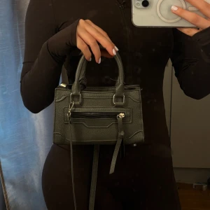 Mörkgrå minihandväska med dragkedja - Säljer en snygg mörkgrå minihandväska i skinn med både handtag och lång axelrem. Väskan har en cool dragkedjedetalj framtill och silverfärgade metalldetaljer. Perfekt för dig som vill ha en stilren och trendig accessoar till din outfit.