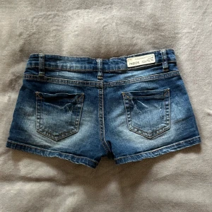 Blå lågmidjade korta jeansshorts från Inside Girl - Snygga blå korta lågmidjade jeansshorts från Inside Girl med klassisk femficksdesign och slitna detaljer. Shortsen har normal passform, låg midja och en söt liten blomdetalj på myntfickan. Perfekta för varma sommardagar och enkla att matcha med allt. Storlek Xs.
