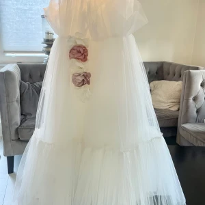 Vit långklänning med volang och blommor - Drömmig vit långklänning för flickor i st 6-8 år  i tyll med volangdetaljer upptill och vackra 3D-blommor i vitt, rosa och lila längs midjan. Klänningen har v-ringad överdel med tunna axelband och markerad midja med knytband baktill. Perfekt för speciella tillfällen.