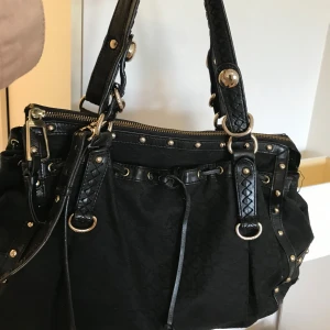 Svart axelväska från DKNY med nitar - Snygg svart axelväska från DKNY med guldfärgade nitar och detaljer. Väskan har flätade handtag, dragkedja upptill och dekorativa ringar. Materialet är skinn och insidan är fodrad med ljust tyg med DKNY-logga. Perfekt för dig som gillar statement-accessoarer.