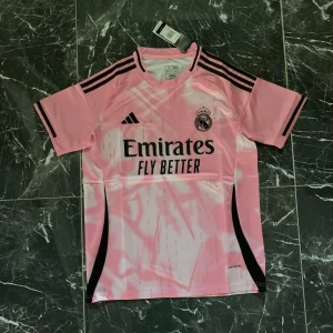 Rosa Real Madrid fotbollströja Adidas - Snygg rosa Real Madrid fotbollströja från Adidas med svarta detaljer och tryck. Tröjan har korta ärmar, svarta ränder på axlarna och klubbmärke på bröstet. Materialet är lätt och andas, perfekt för träning eller match. Emirates Fly Better-logga på framsidan.