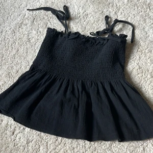 Svart smockad linnetopp från Loness - Supersöt svart linnetopp från Loness med smockad överdel och volang nertill. Axelbanden är tunna och knyts på axlarna för en extra gullig touch. Perfekt till jeans eller kjol när du vill ha en chill men ändå snygg look. Kan även användas som kjol🥰