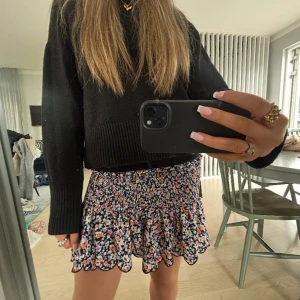 Blommig kortkjol med volangkant - Supersöt kortkjol med blommigt mönster i svart, vit, orange, rosa och lila. Kjolen har smockad midja och volangig nederkant med vågig avslutning. Perfekt för dig som gillar färg och vill ha en söt look❤️